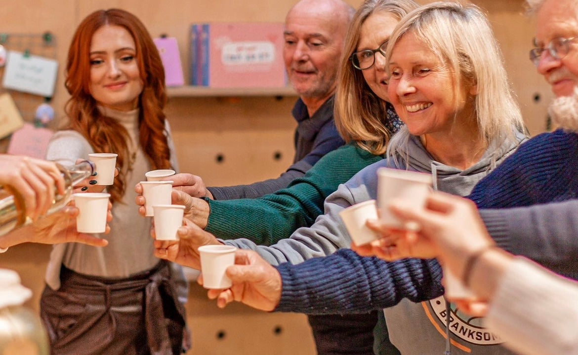 Photo of kombucha workshop students "cheers to our gut health" with small glasses of delicious looking kombucha, to conclude a session, happy smiley faces of all ages with our proud founder Hebe, pouring a sample for herself - boochacha