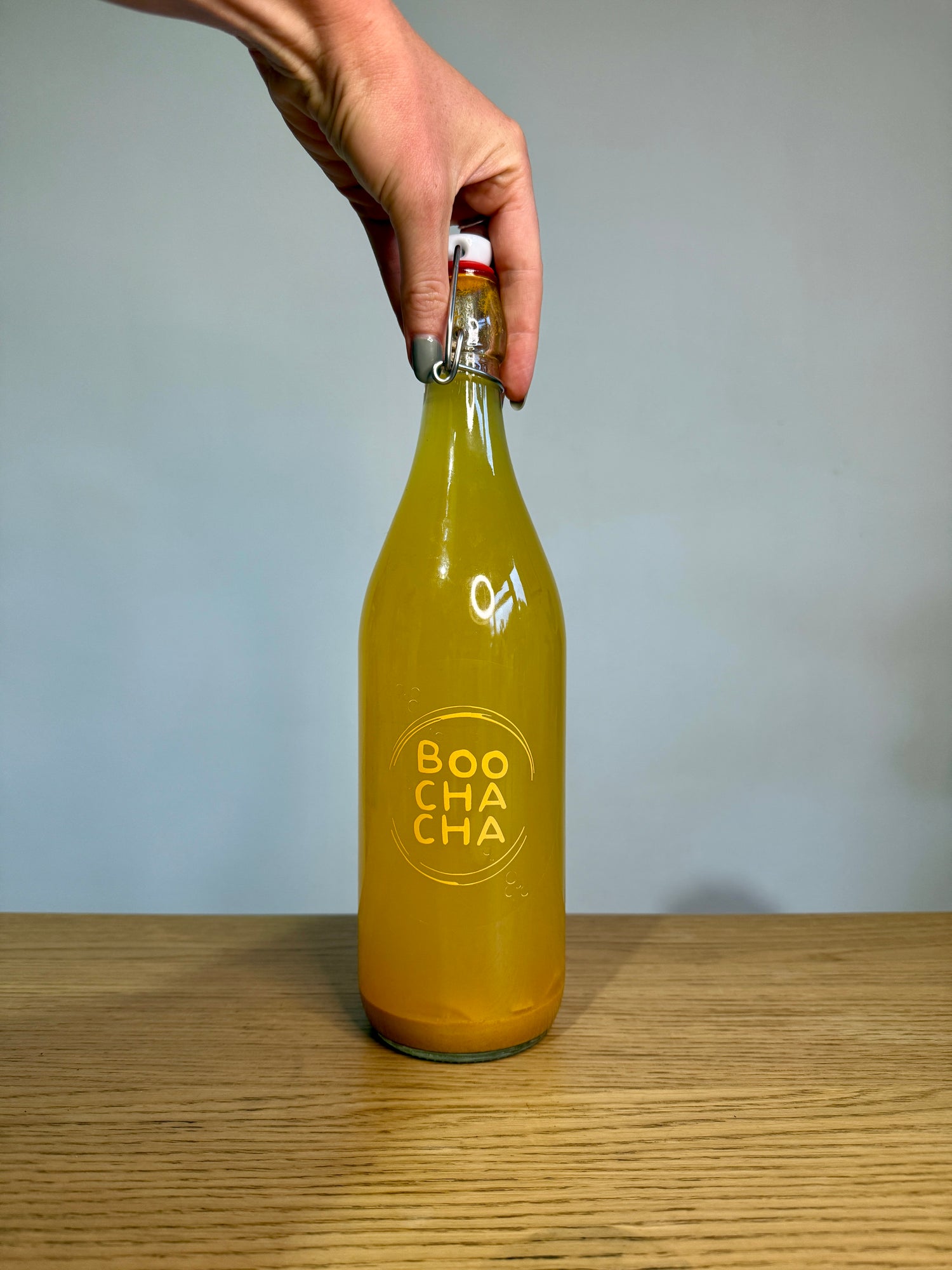 Ladies hand holding a bottle of homemade ginger kombucha (Boochacha) on a wooden surface with a grey background